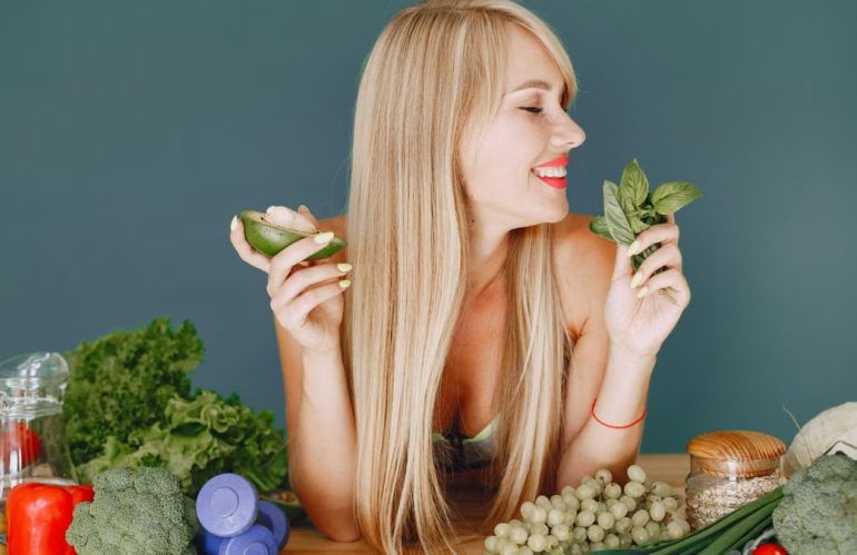 A smiling woman holding avocado and basil, surrounded by fresh vegetables and fruits for healthy eating.